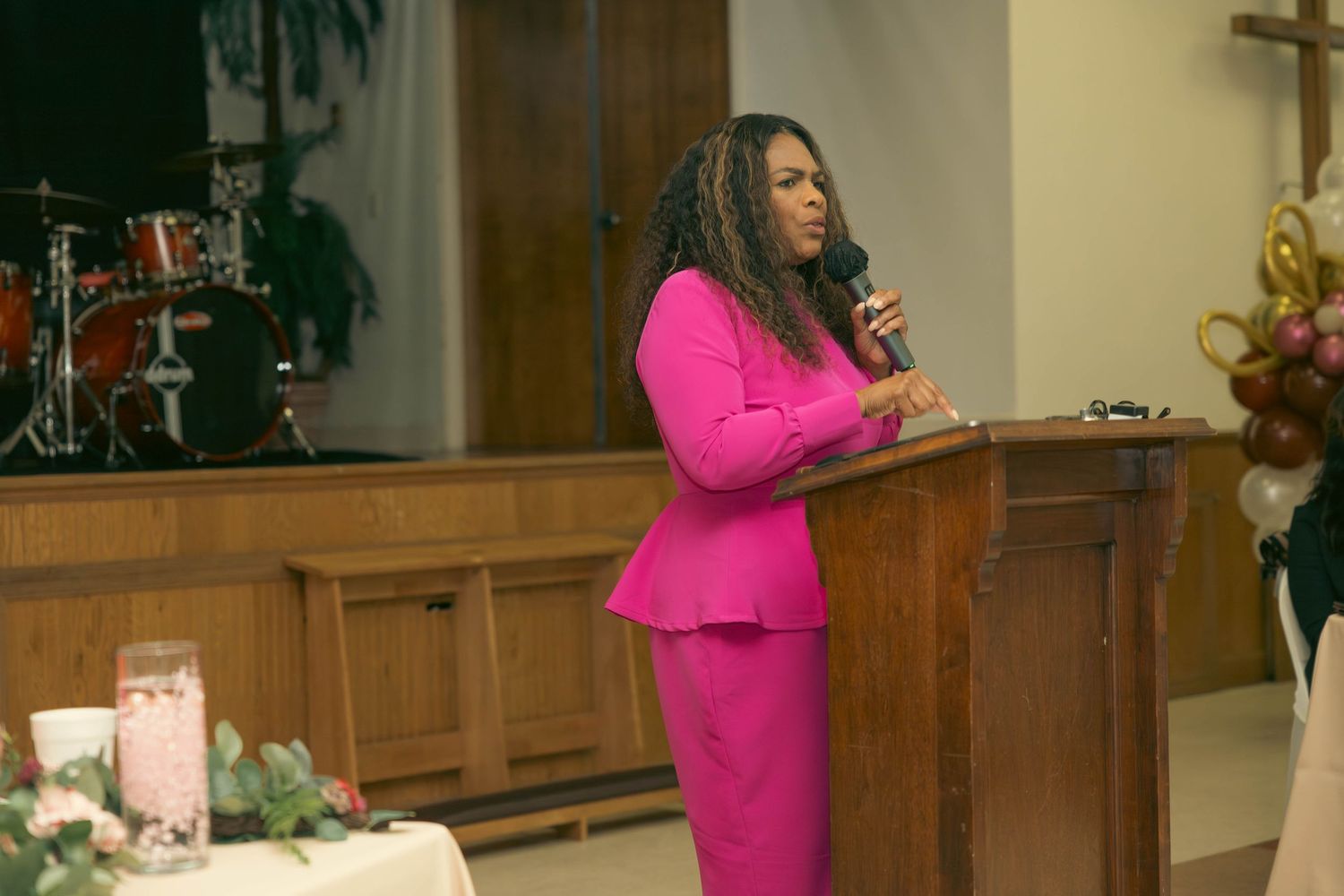 Woman in bright pink suit speaking at a podium during a church service or ceremony.