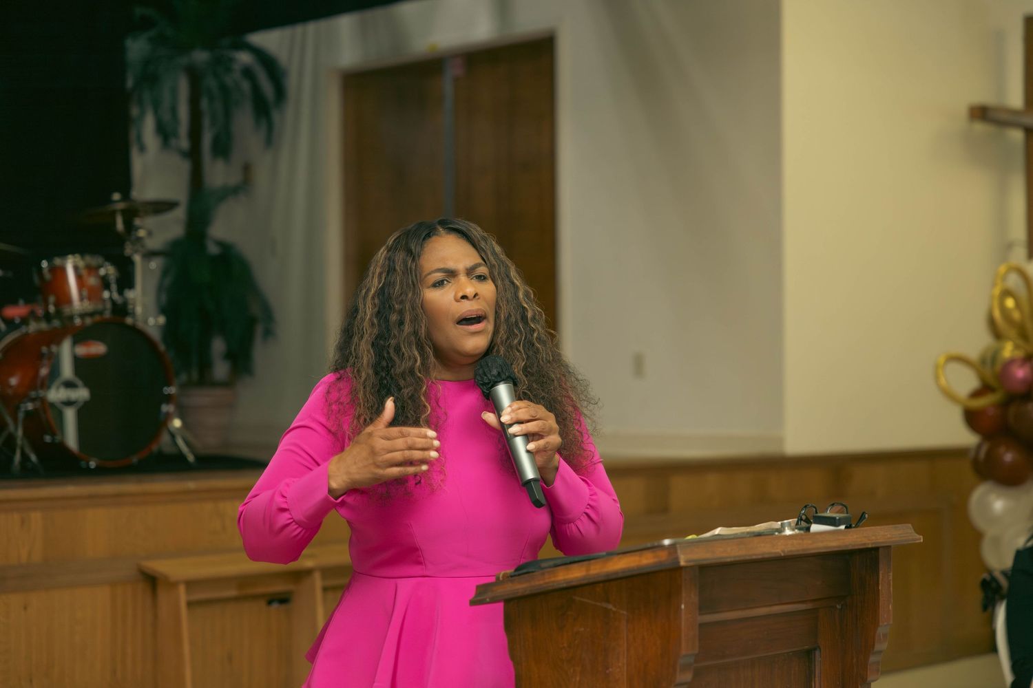 Woman in pink dress passionately singing into microphone at a church event near podium.