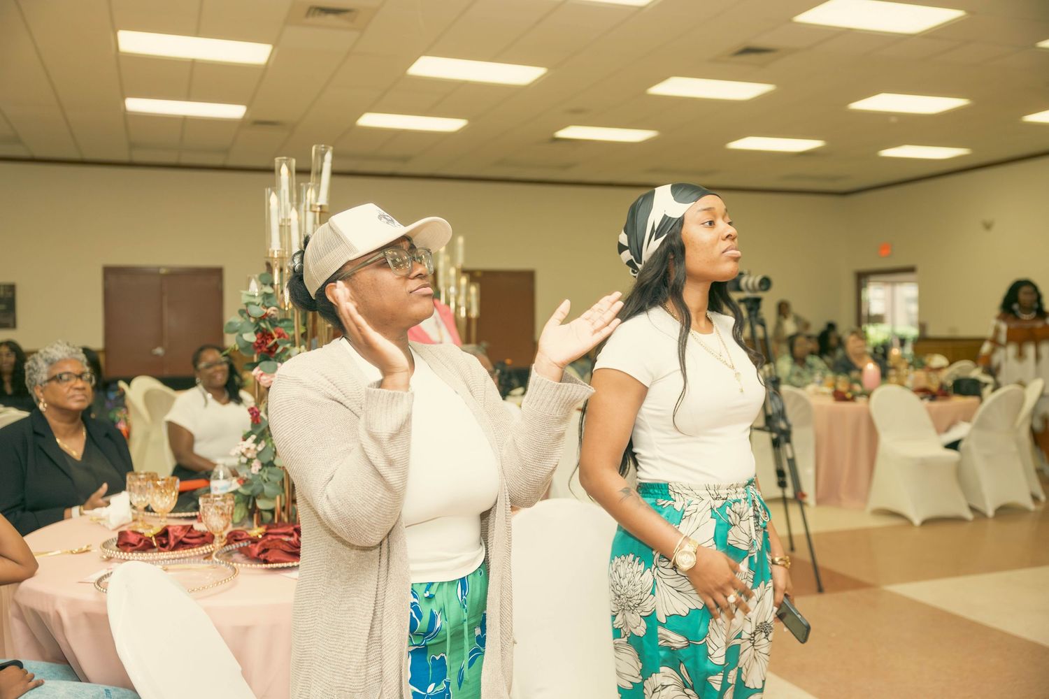 Two women in elegant hats worship during a church event in a decorated banquet hall.