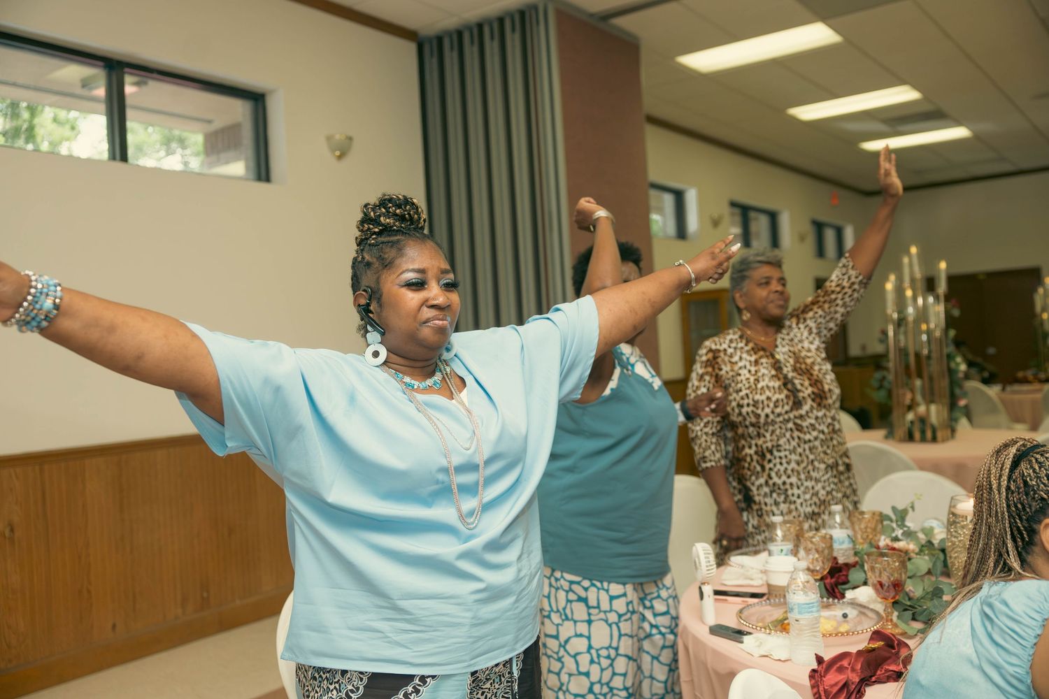 Women raise their arms in praise and worship during a church gathering in a decorated hall.