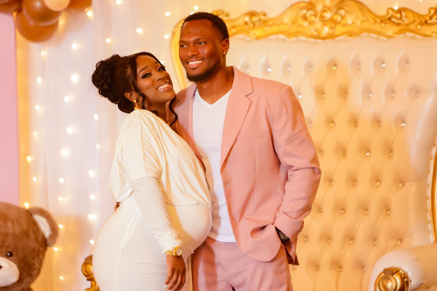 Pregnant woman in white dress poses with man in pink suit at elegant baby shower.