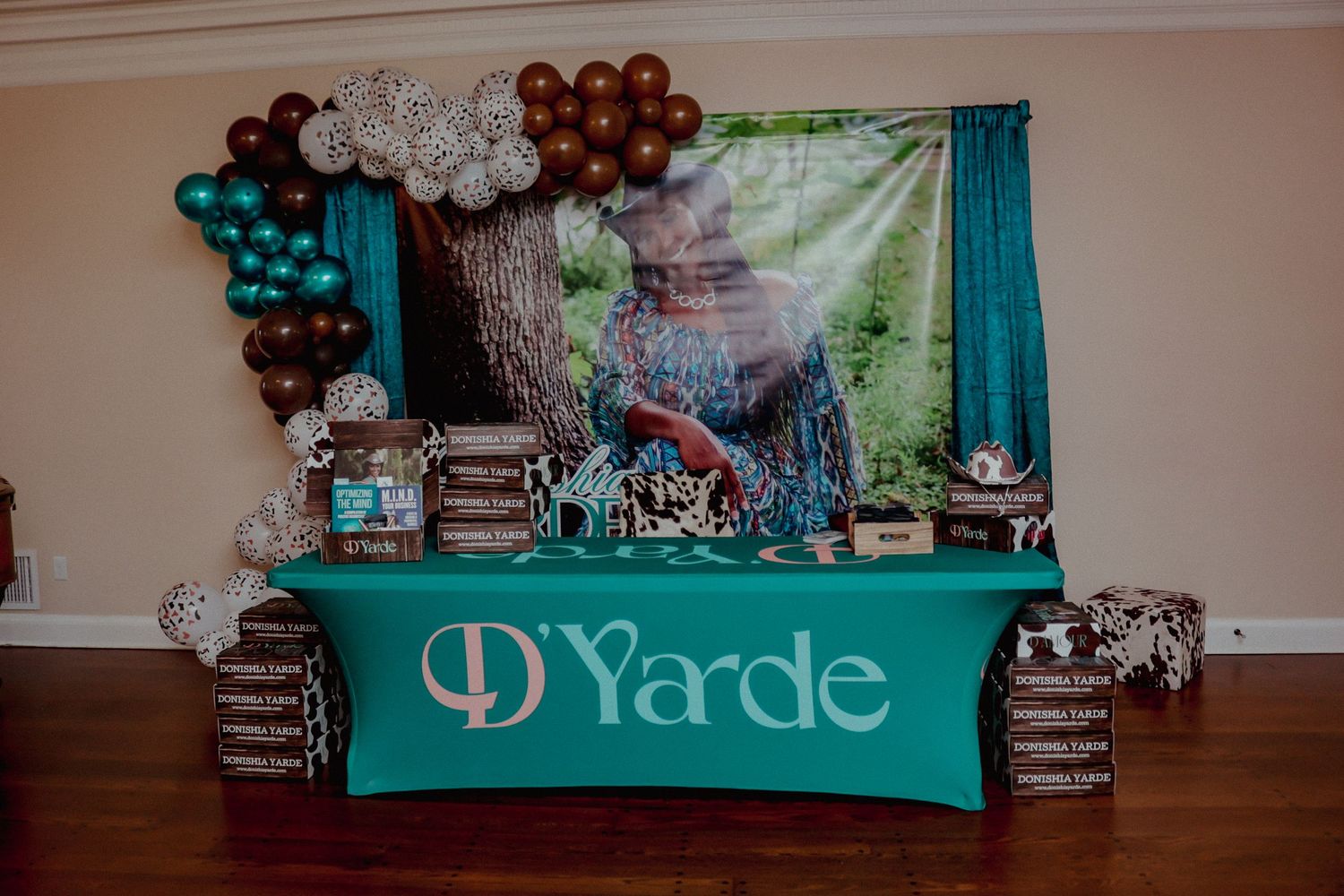 D'Yarde branded teal tablecloth display with balloon garland and photo backdrop.