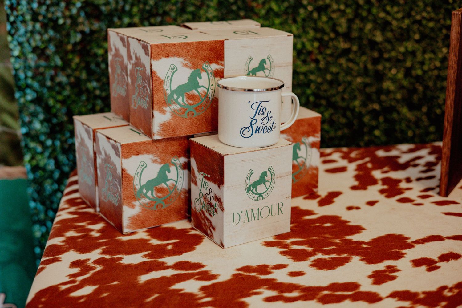 Decorative gift boxes with cow print and branded mugs stacked on a patterned table.