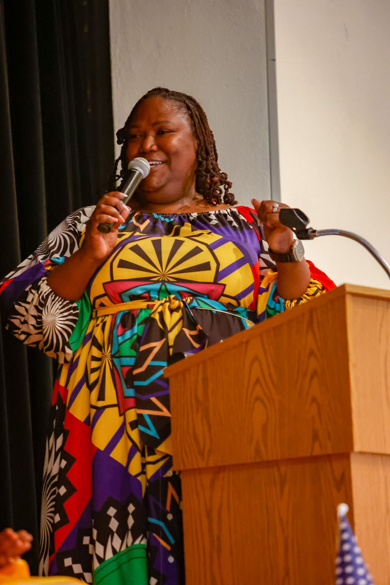 Woman in colorful African print dress speaks passionately into microphone at wooden podium during school event.