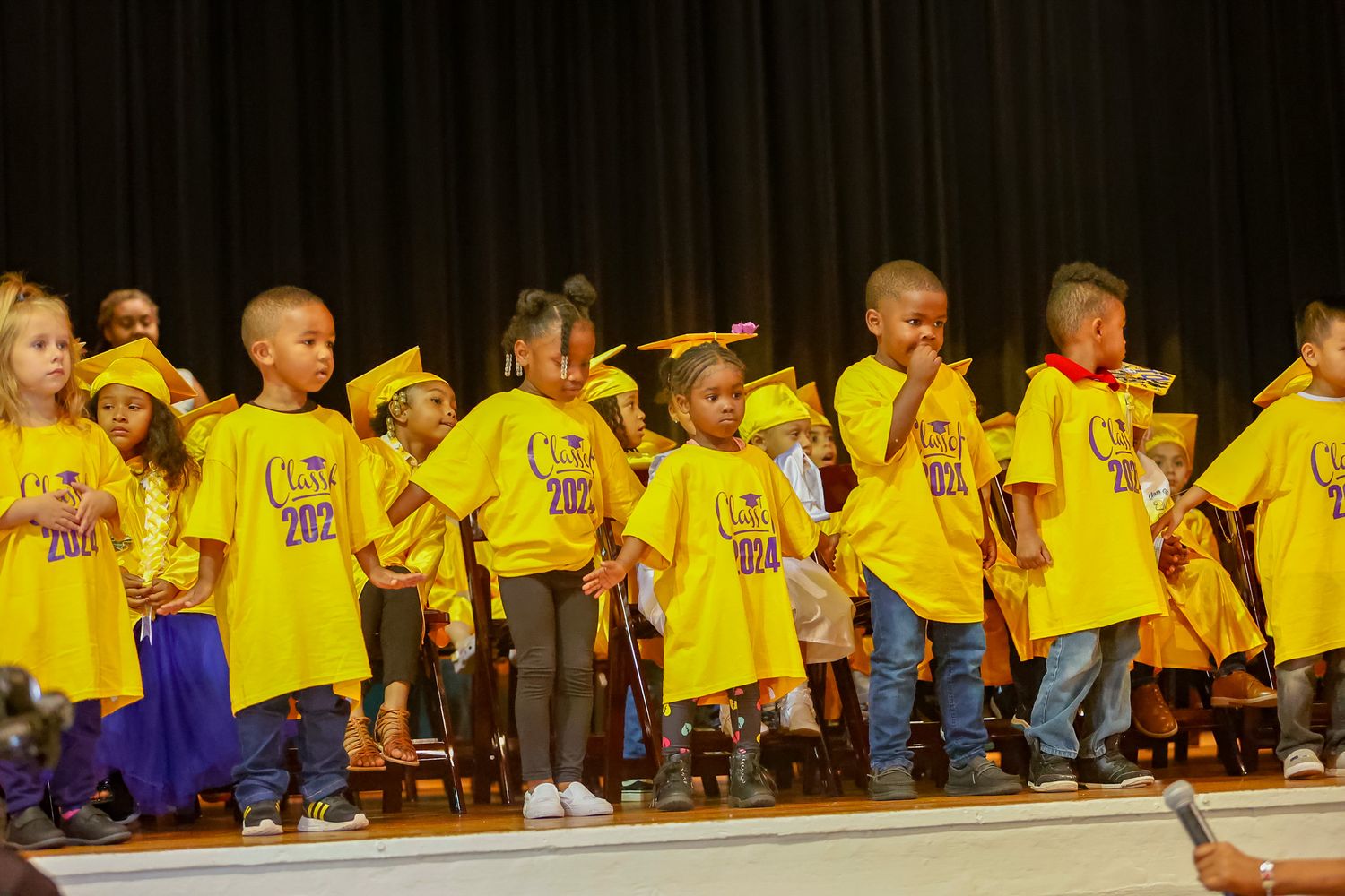 Young children in yellow graduation caps and gowns perform on stage during preschool graduation ceremony.