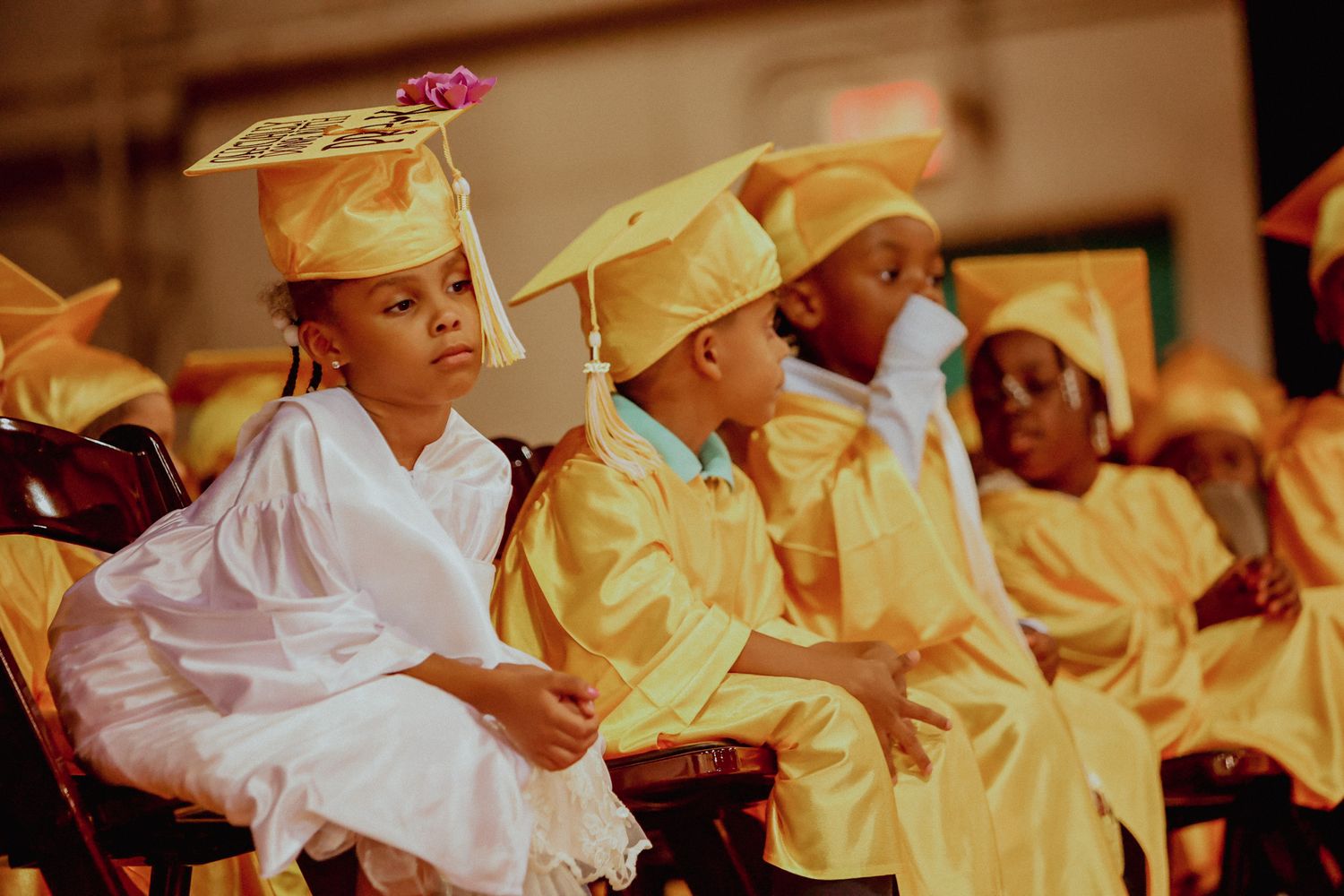 Young children in gold graduation caps and gowns sit in chairs during preschool graduation ceremony.