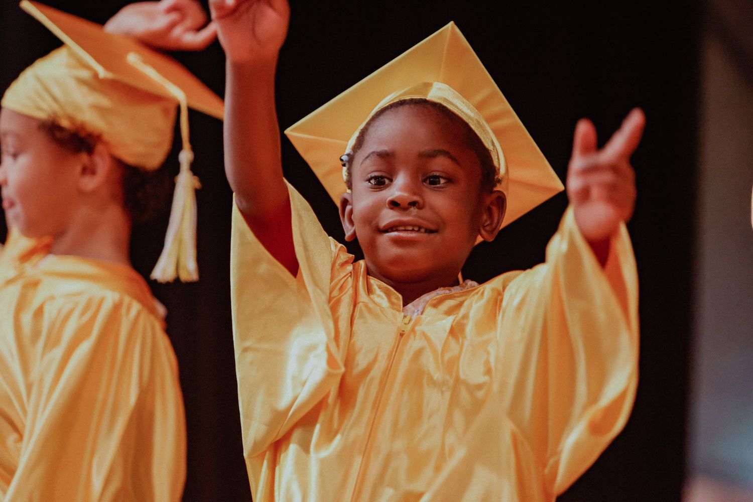 Young child in gold graduation cap and gown raises hand proudly during preschool graduation ceremony.