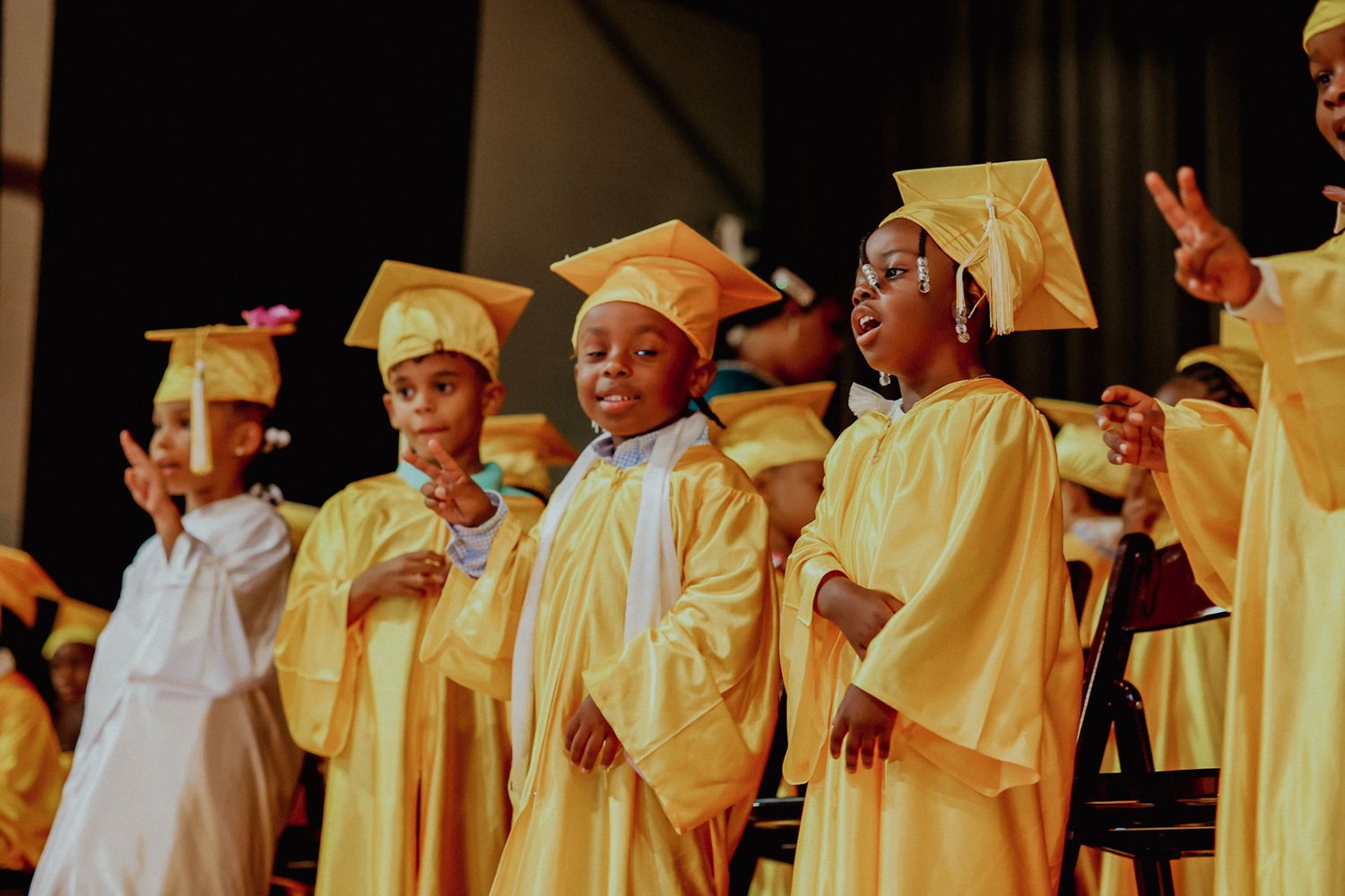 Young children in gold graduation caps and gowns perform and gesture on stage at preschool graduation.