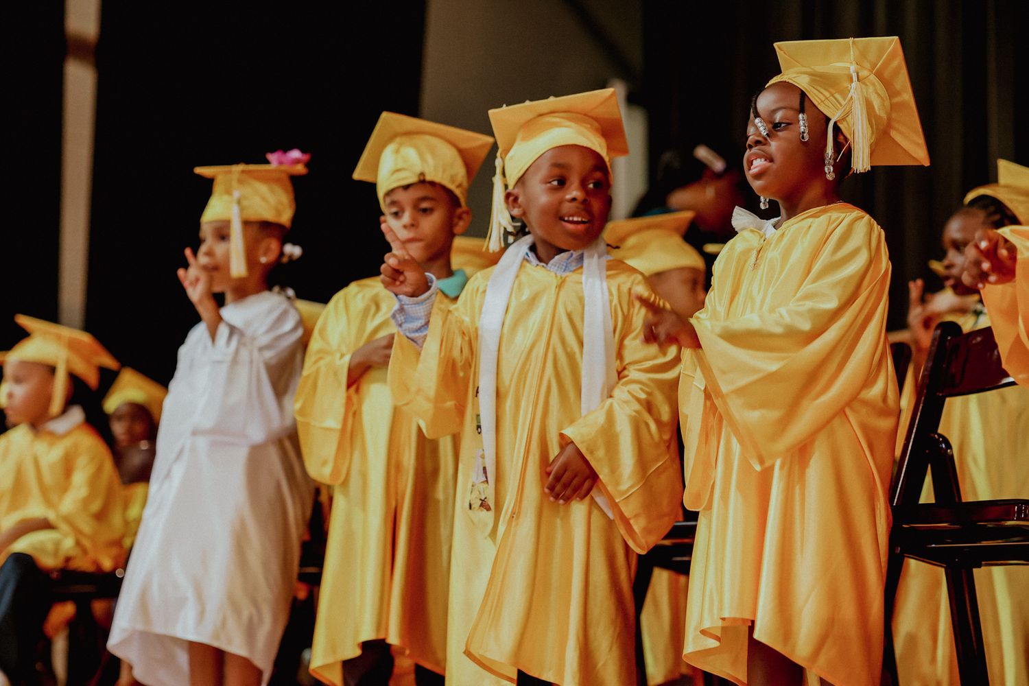 Three young children in gold graduation gowns stand on stage performing at preschool graduation ceremony.