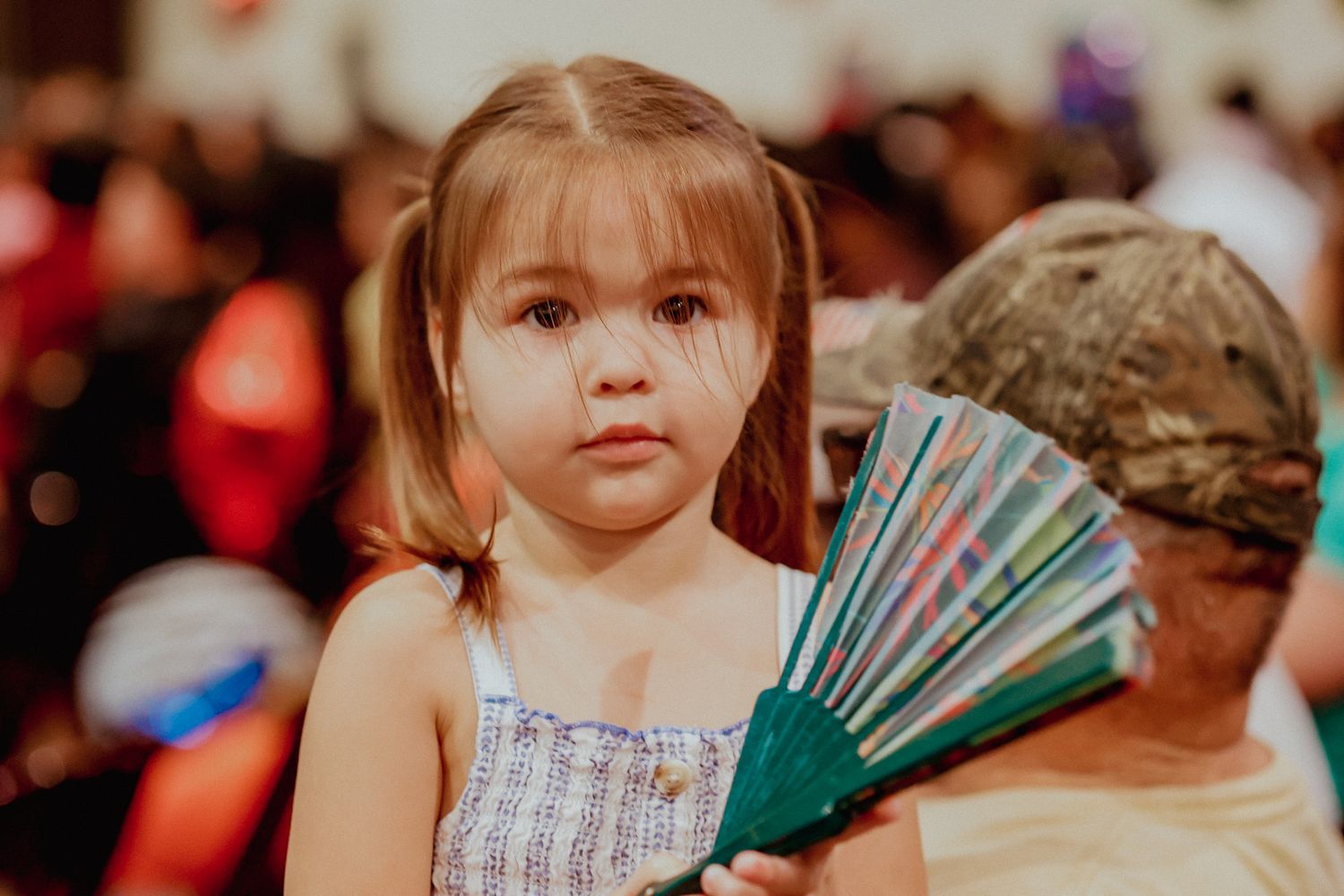 Young girl with pigtails holding a colorful hand fan at an indoor event.