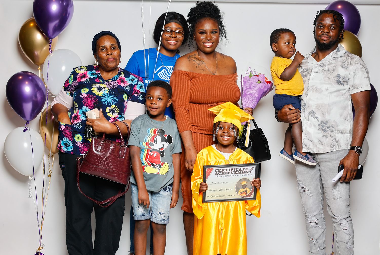 Family group celebrating a childs kindergarten graduation with balloons and a certificate indoors.