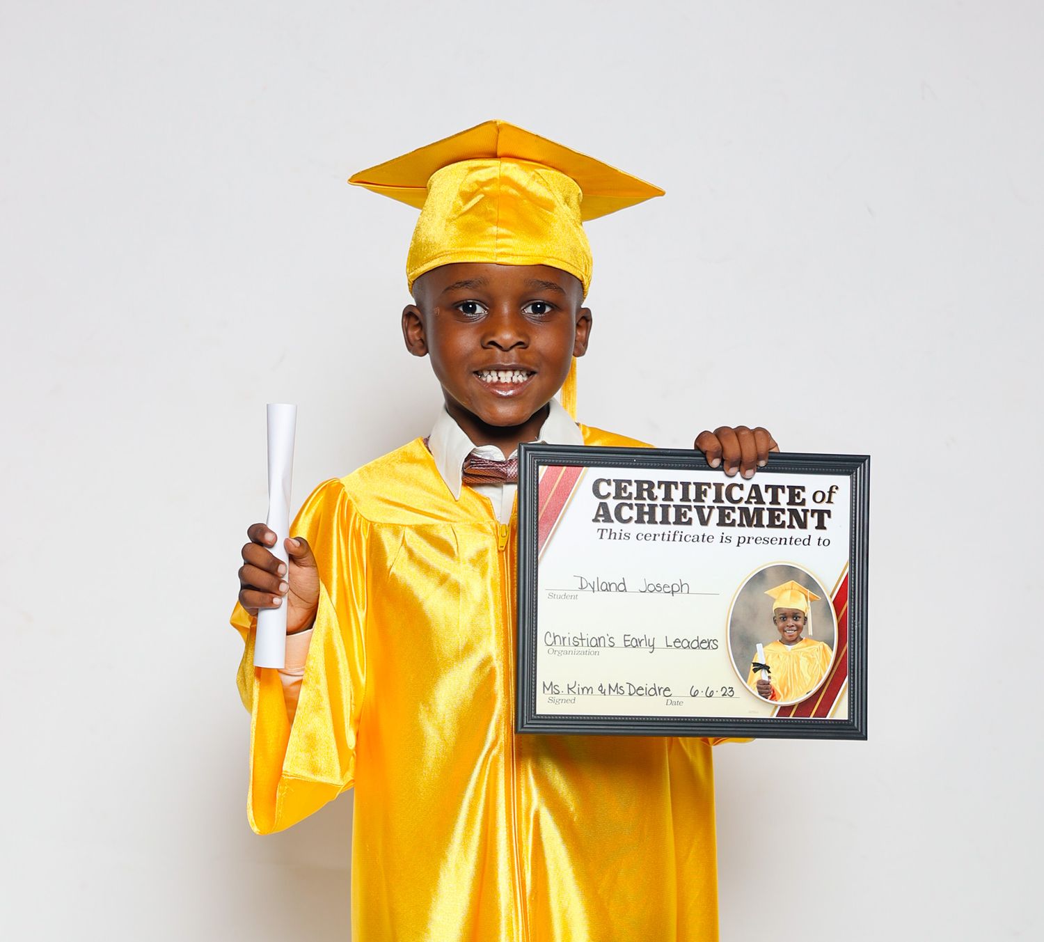 Young boy in yellow graduation cap and gown proudly holding a Certificate of Achievement and diploma.