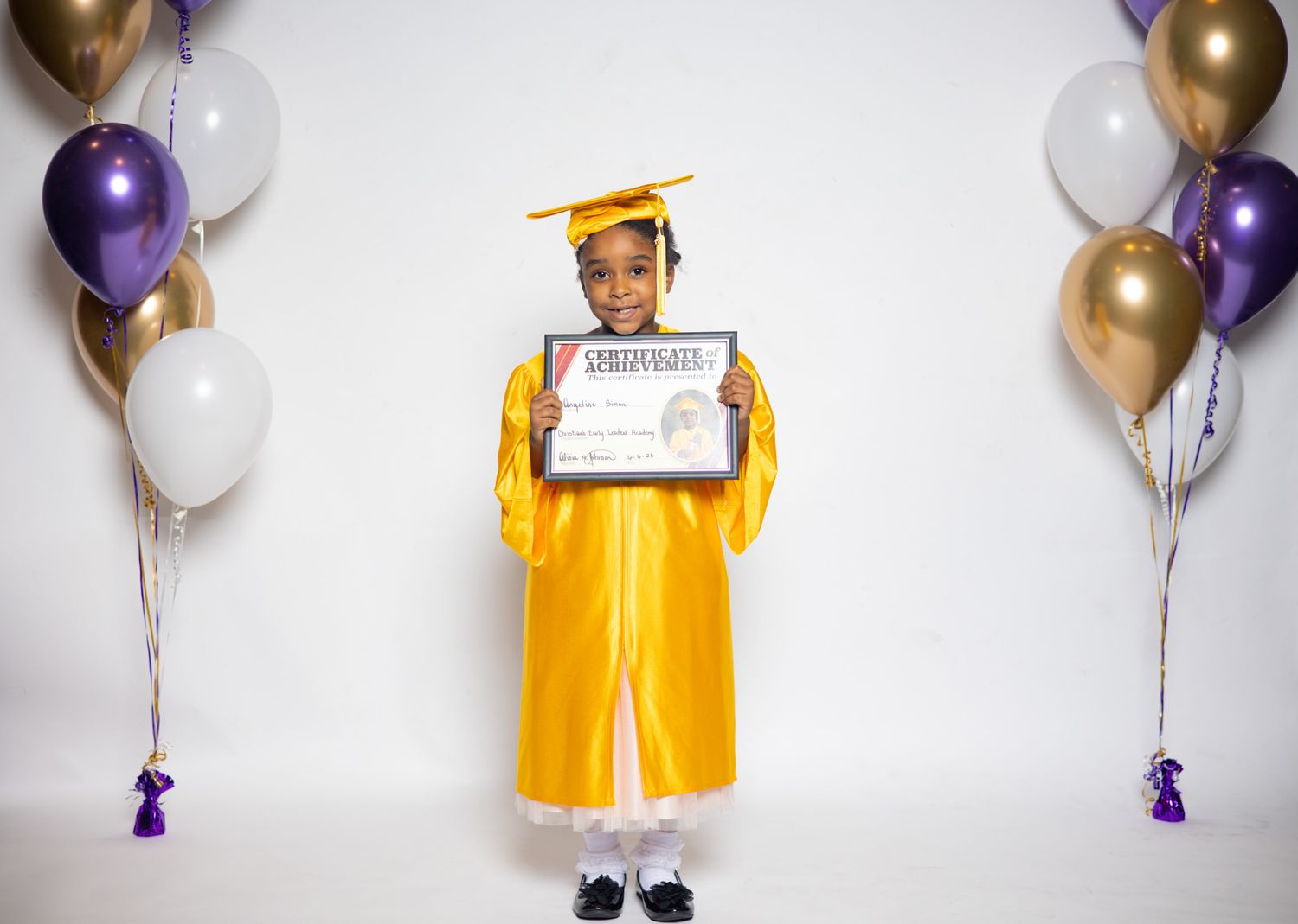 Child in yellow graduation gown holding certificate surrounded by purple, gold, and white balloons.