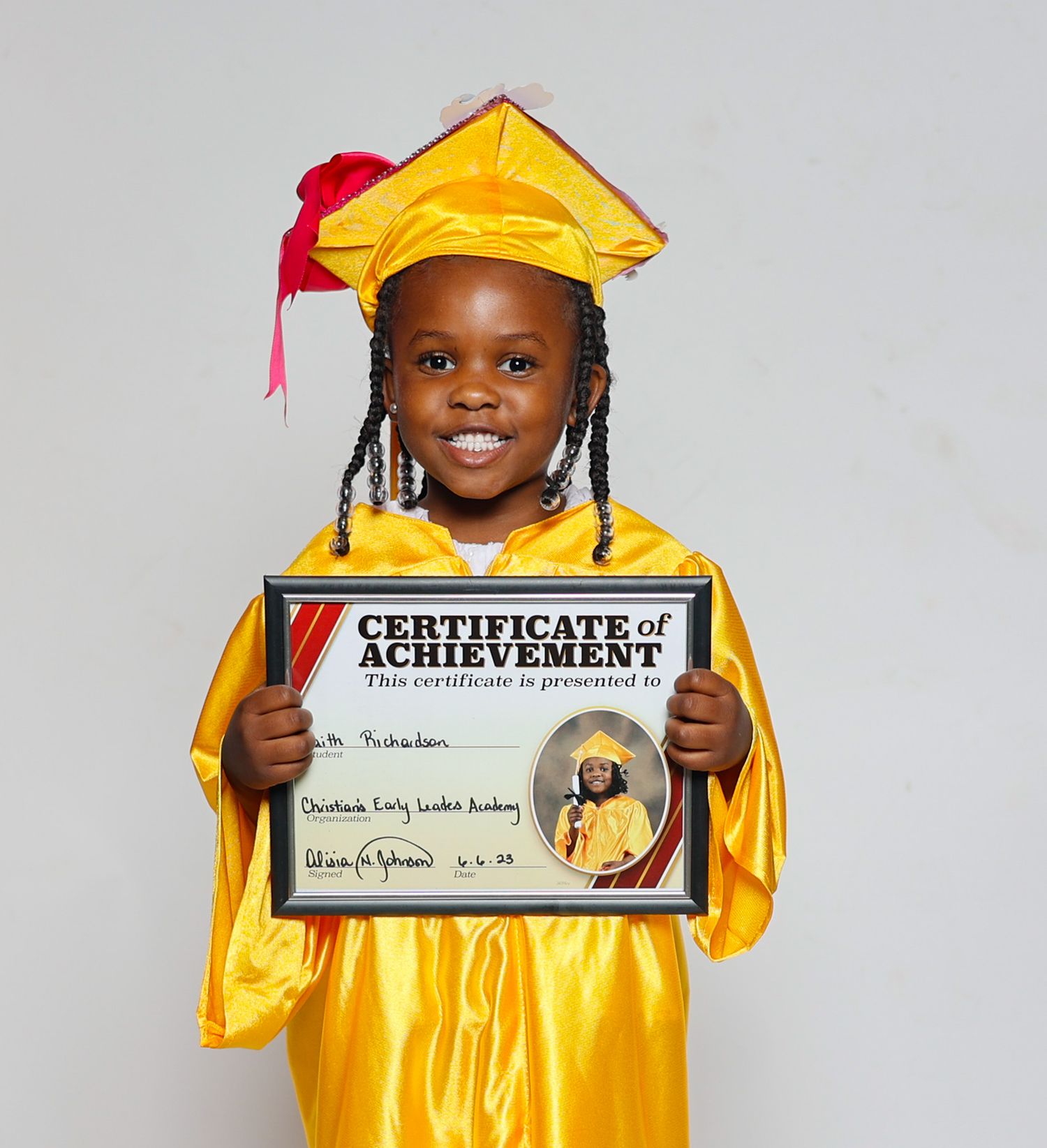 Smiling young girl in yellow graduation cap and gown proudly displaying her Certificate of Achievement.