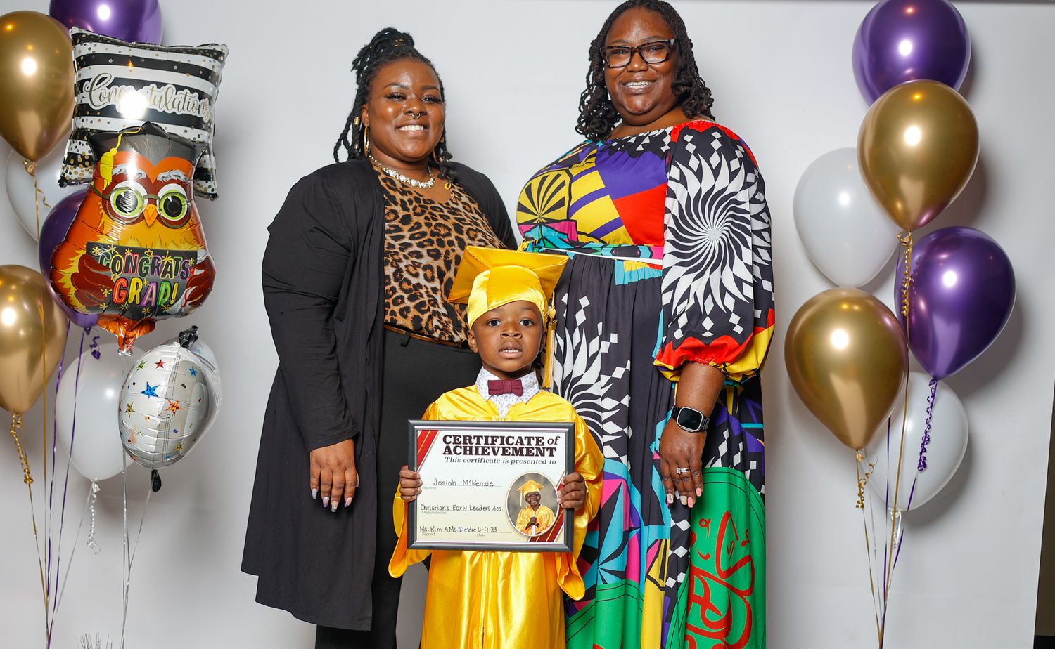 Young graduate in gold cap and gown holds diploma with two proud family members at graduation ceremony with colorful balloons.