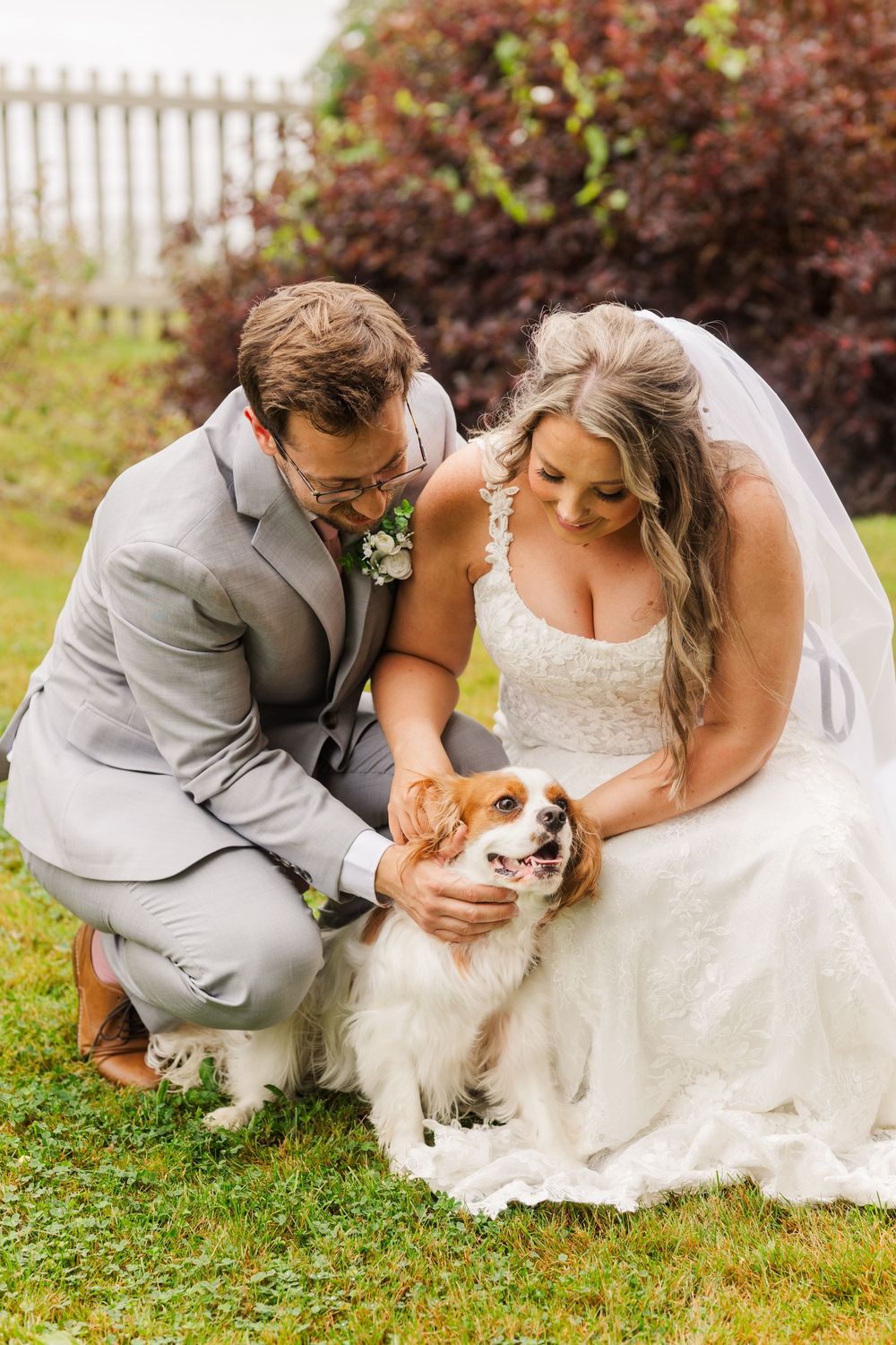A bride and groom in formal wedding attire share a tender moment on grass with their small white dog.