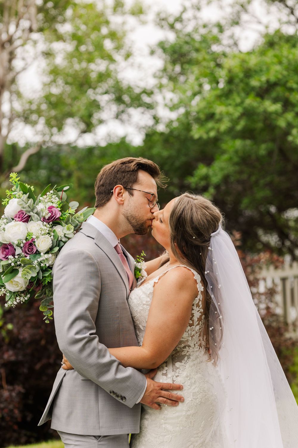 A newlywed couple shares intimate kisses in their wedding attire outdoors with lush greenery in the background.