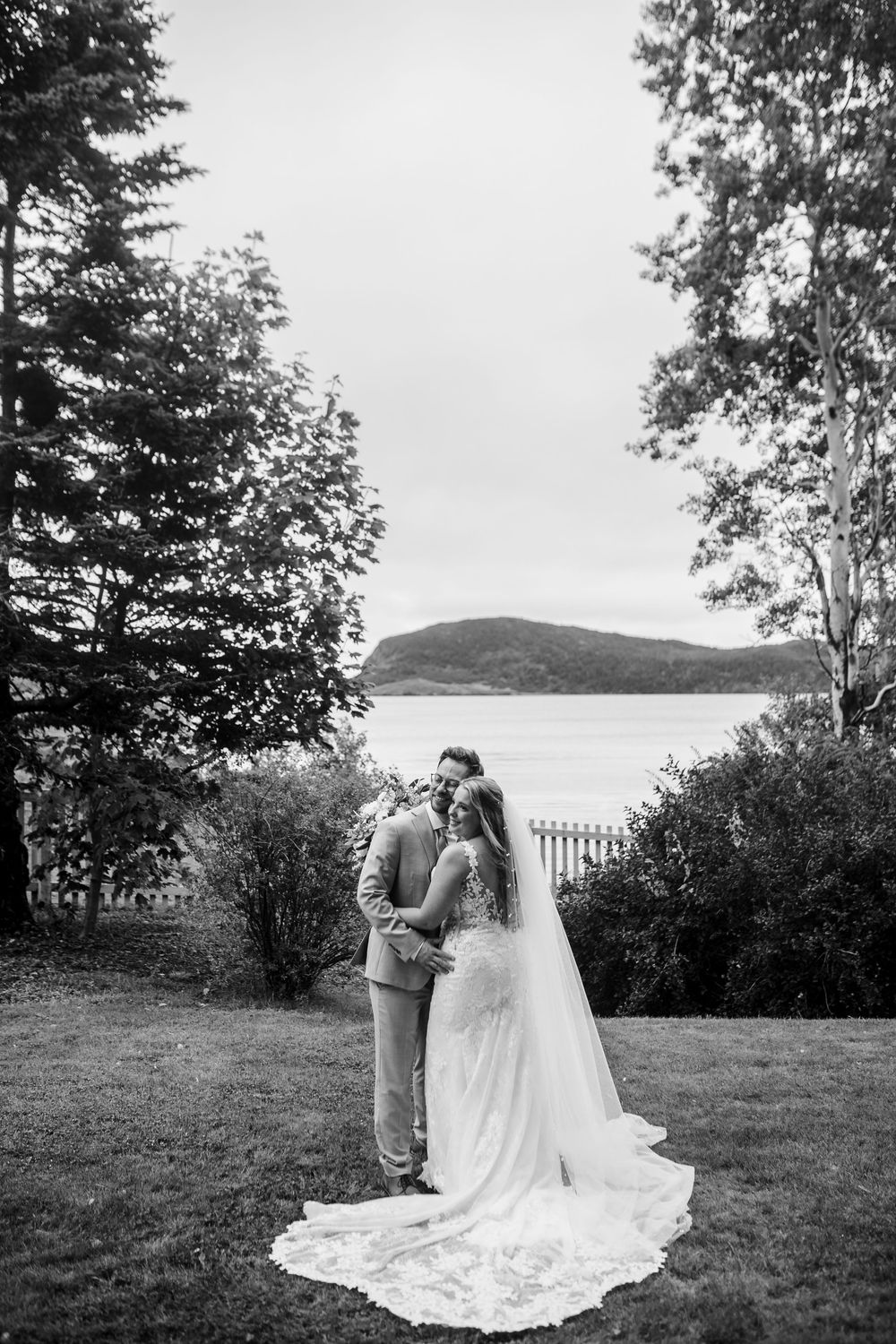 A beautiful long white wedding dress train flows across a grassy lawn with trees and water in the background.