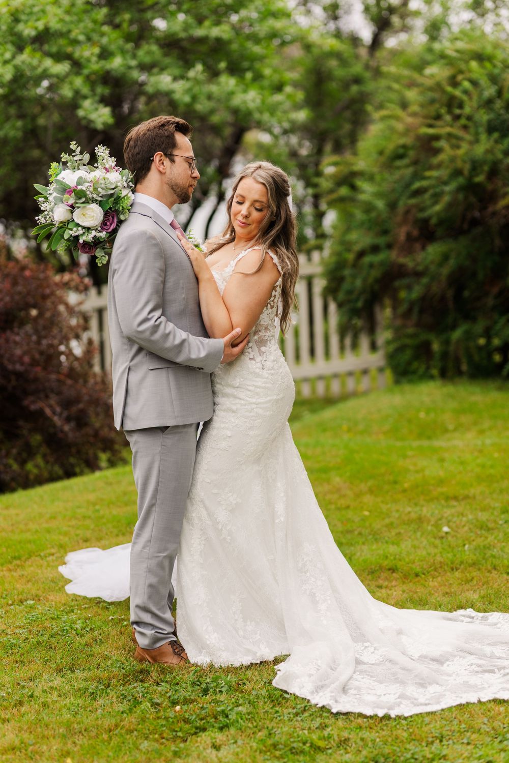 A newlywed couple embraces while dancing on a lush green lawn with white floral decorations in the background.