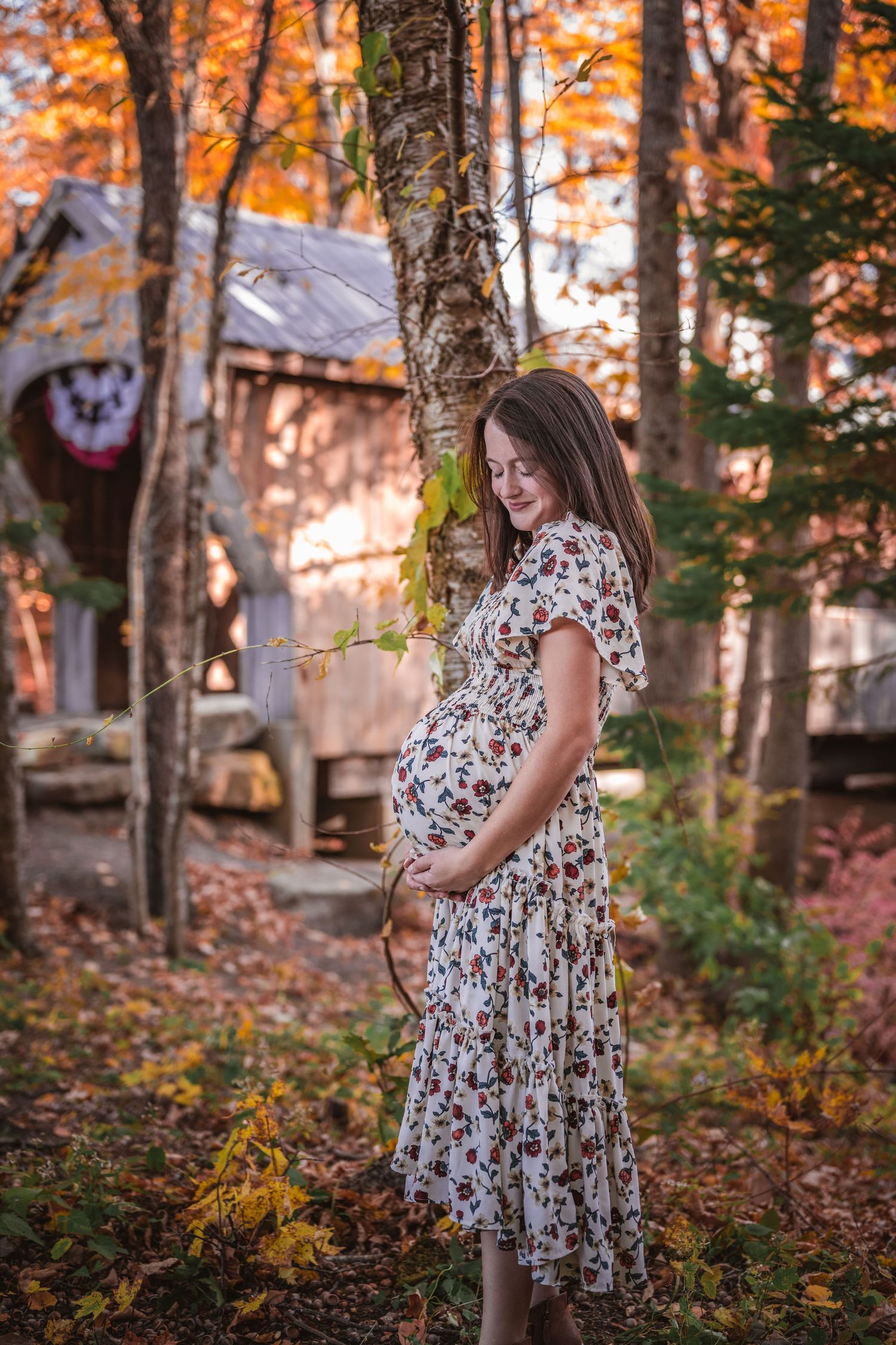 Smiling pregnant woman in floral dress standing among colorful autumn trees near a covered bridge.