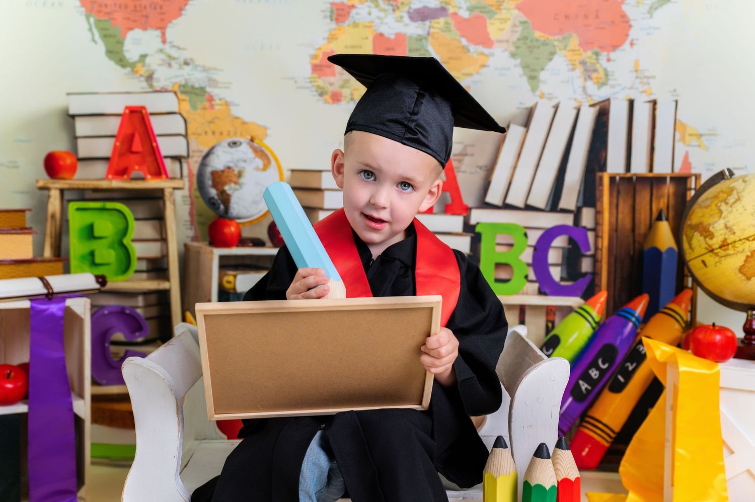 Small student in graduation cap and gown holds up diploma while surrounded by educational toys and supplies.