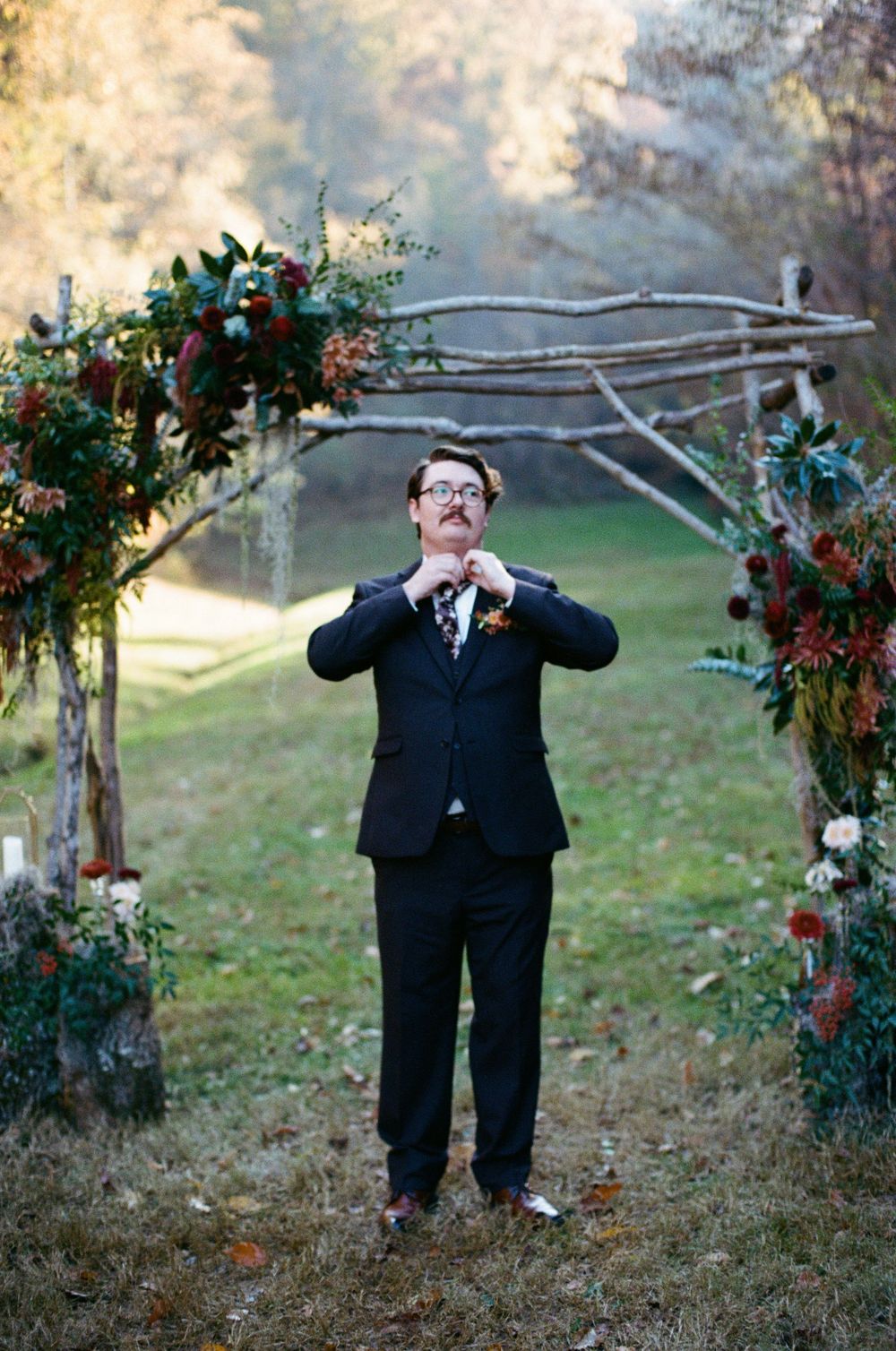 Person in formal attire posing playfully with tree branches at an outdoor wedding setup.