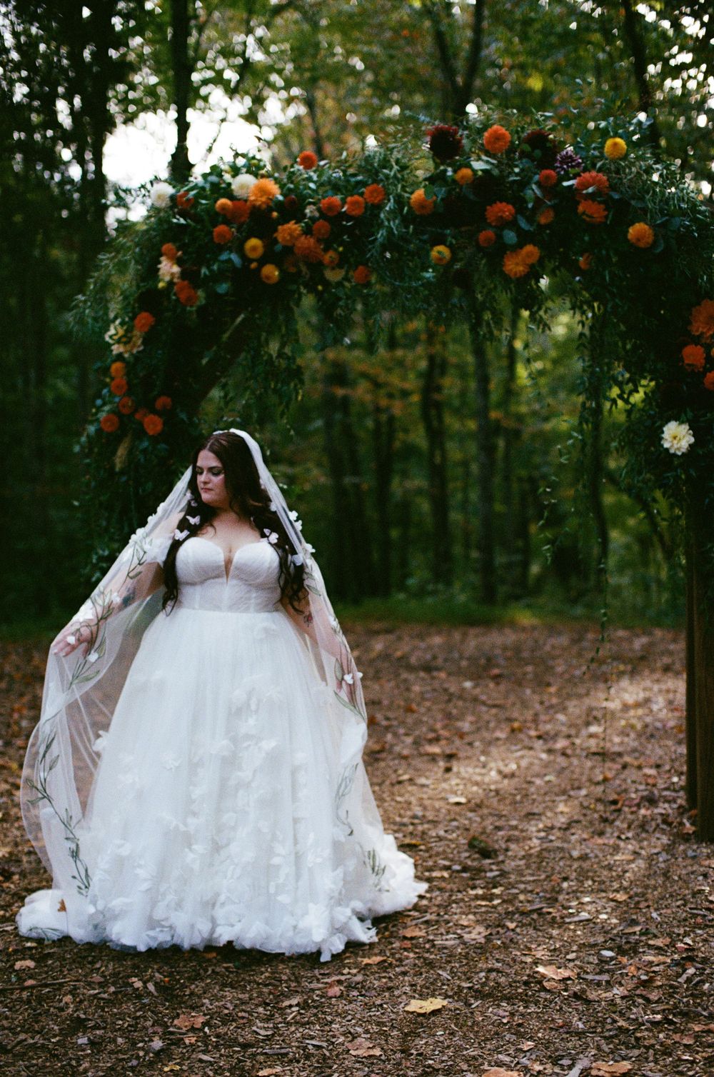 Wedding dress silhouette photographed under orange floral arch in wooded setting.