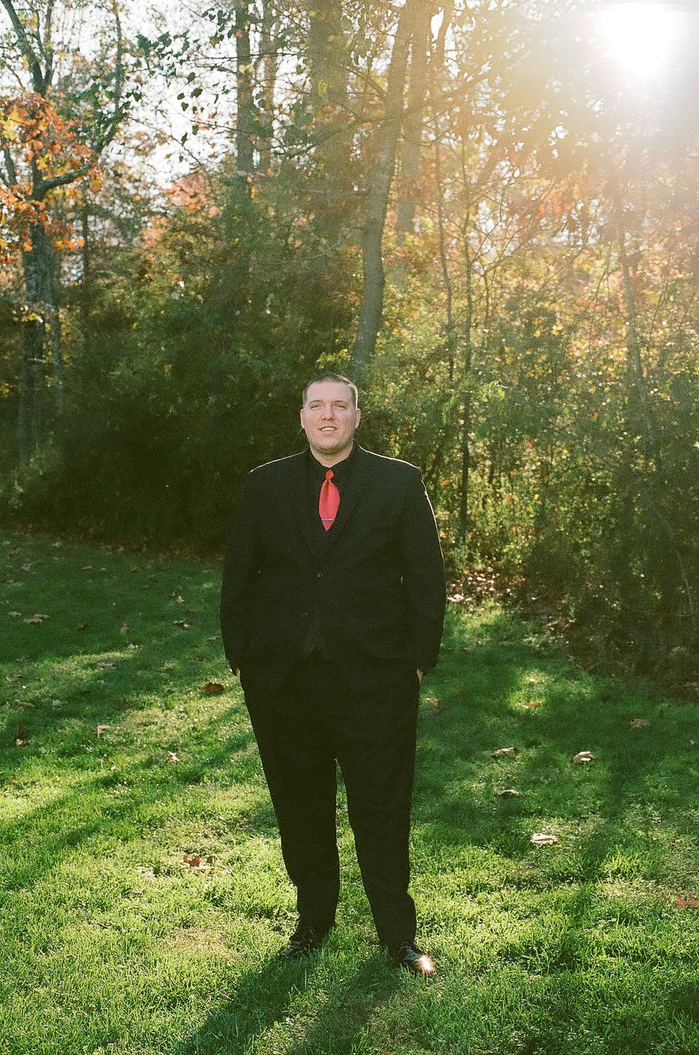 A person in formal black suit and red tie stands in a sunlit autumn garden.