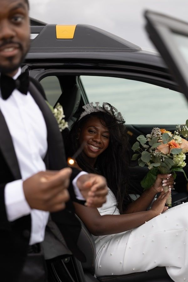 A joyful bride wearing a tiara and holding a floral bouquet sits in a car during a wedding celebration.