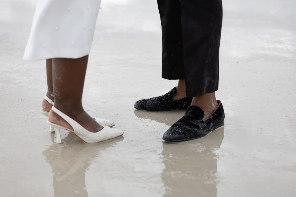 Black formal shoes and white high heels stand together on a reflective surface showing details of their footwear.