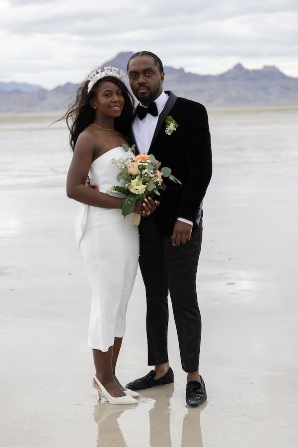 A couple embraces on a salt flat desert landscape while dressed in formal wedding attire with a bouquet.