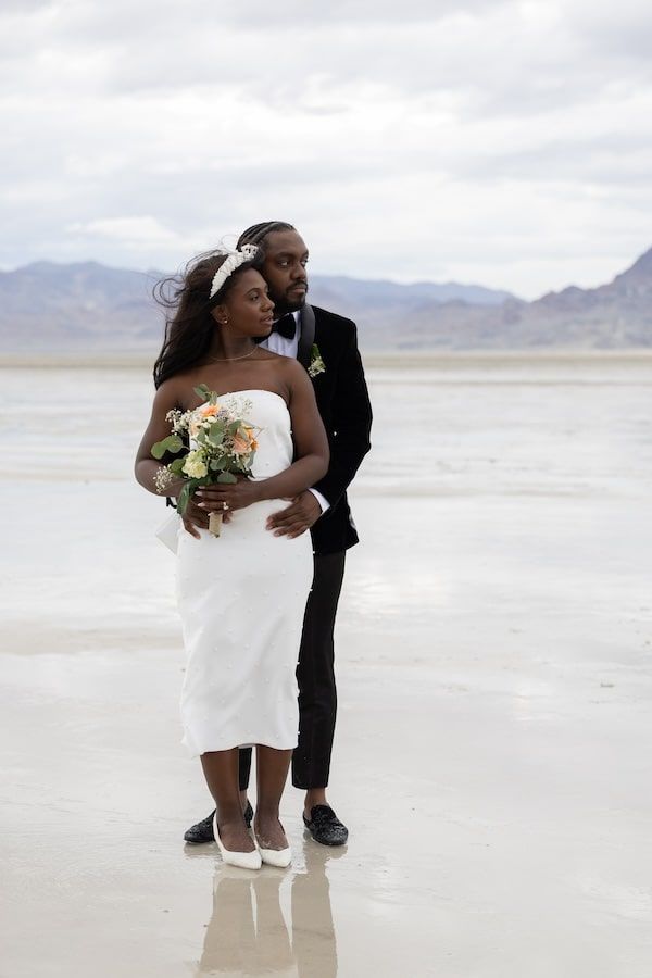 Romantic couple embraces in white wedding attire while standing on bright salt flats with mountains in the distance.