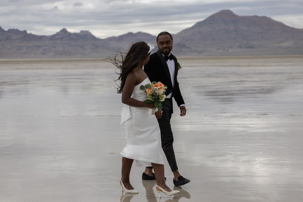 A newlywed couple runs and dances across the salt flats during their wedding photo shoot under dramatic cloudy skies.