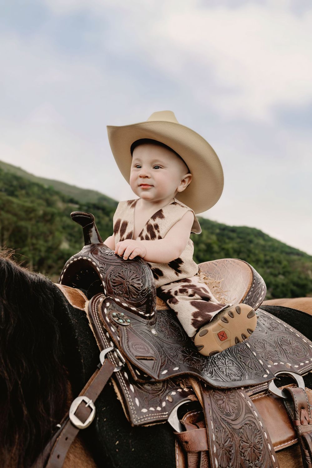 Young rider in cowboy hat sits atop a western leather saddle decorated with ornate tooling against green hillside.