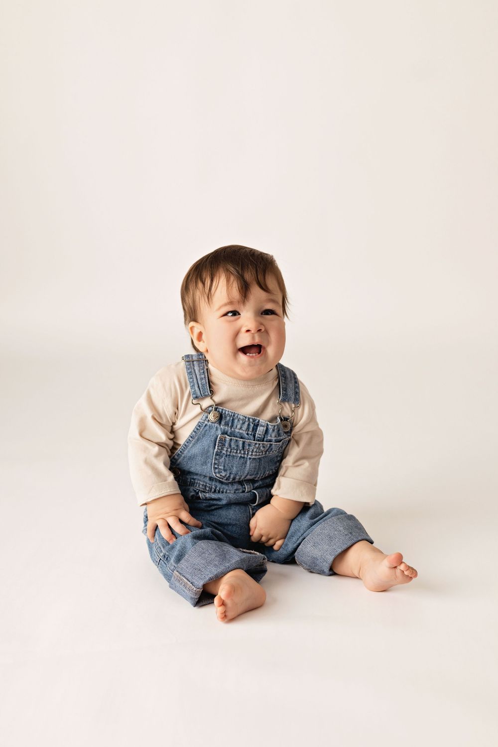 A joyful baby wearing denim overalls and cream shirt sitting and laughing against a white backdrop.
