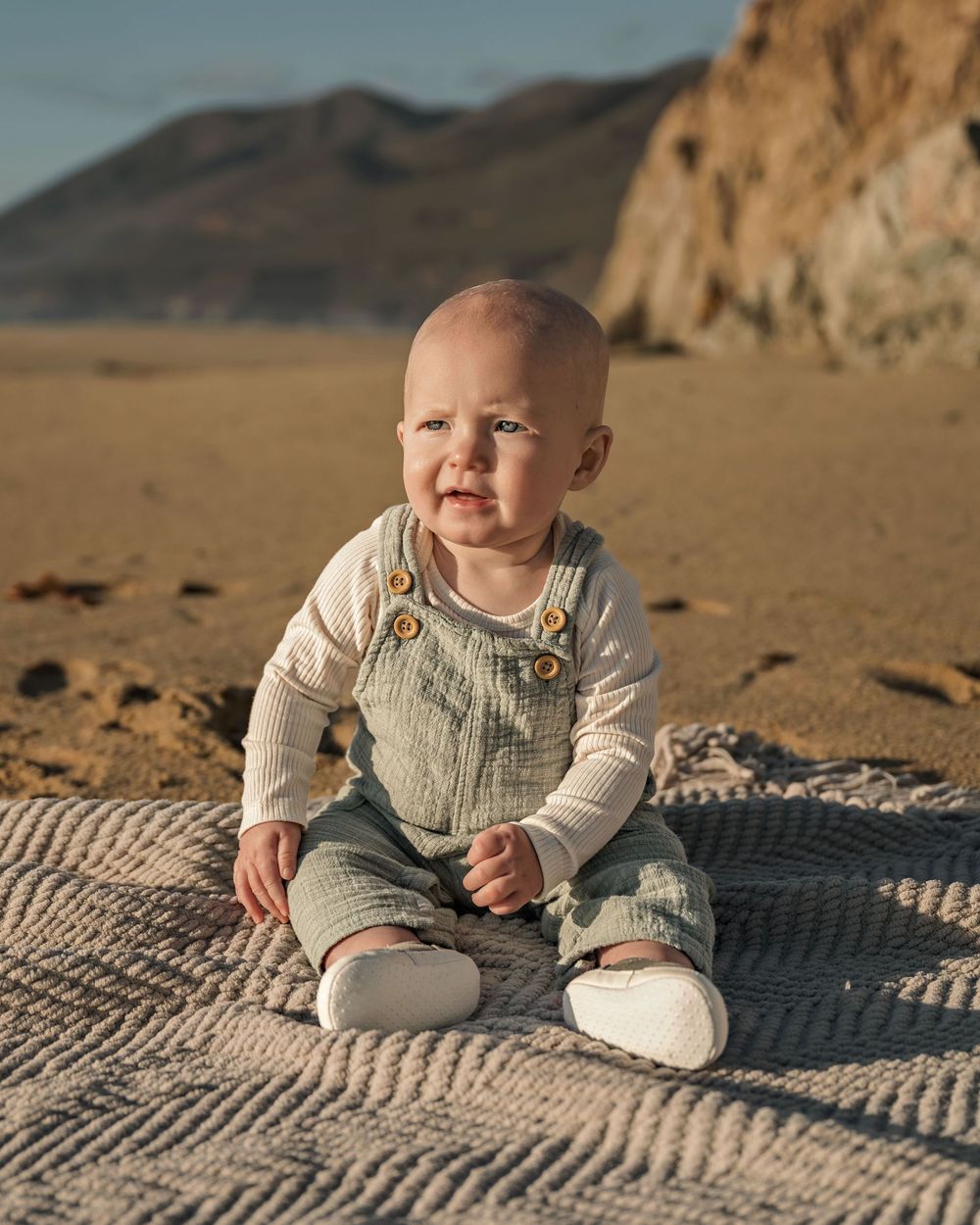 Baby in neutral outfit sitting on sandy beach with cliffs in background.