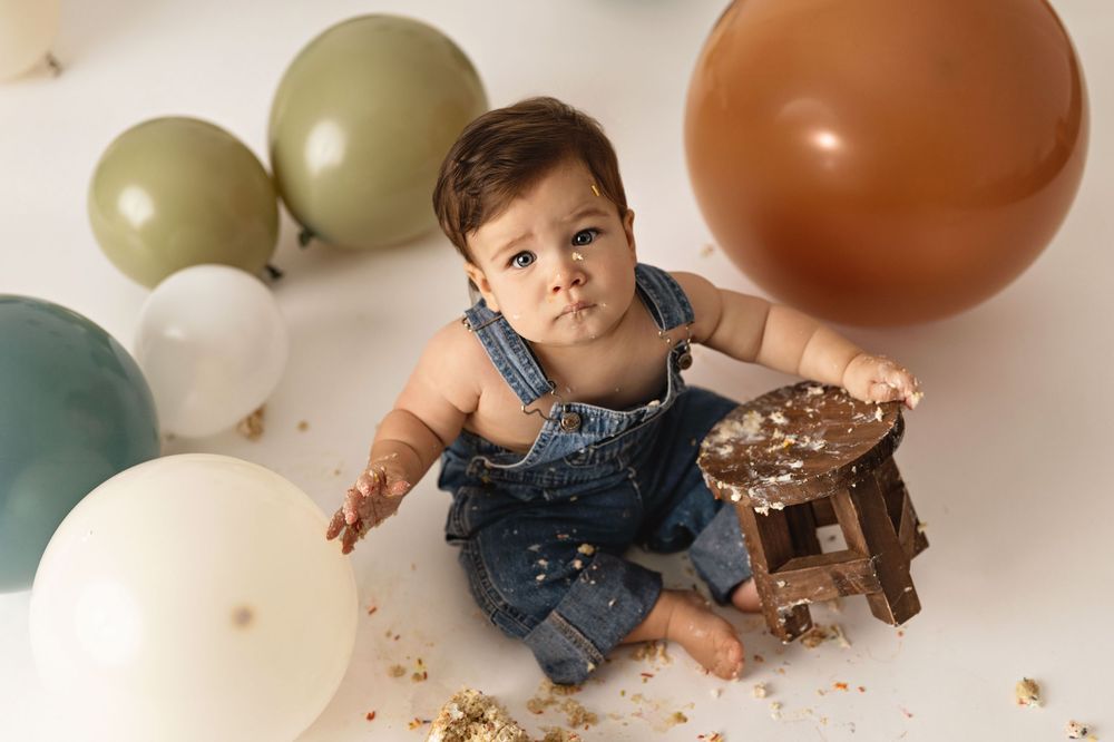 A baby wearing denim overalls plays with cake and balloons during their first birthday smash cake photoshoot.