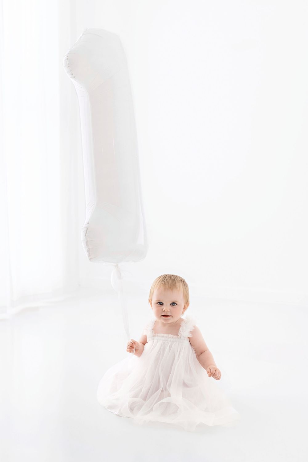 Baby in white dress crawls on white backdrop during minimalist studio photo session.