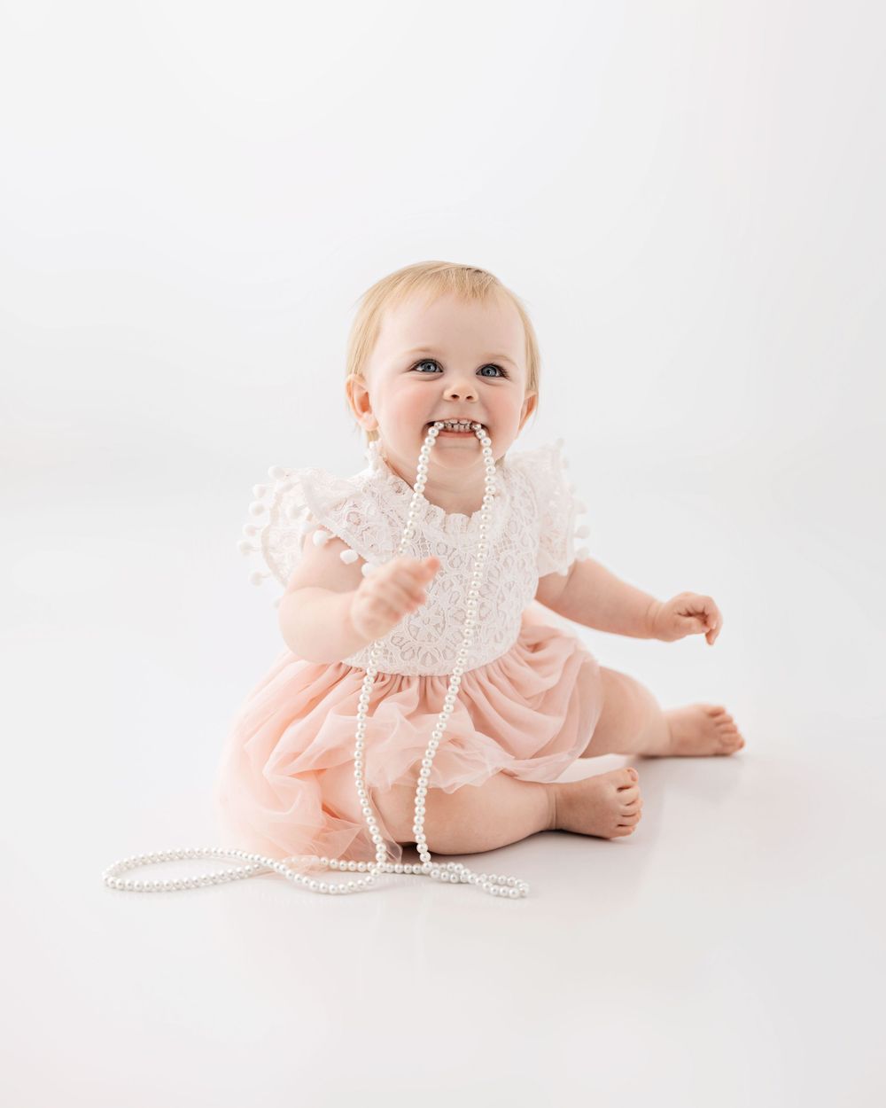 A baby in a pink dress plays with a string of pearls on a white backdrop during a studio photoshoot.