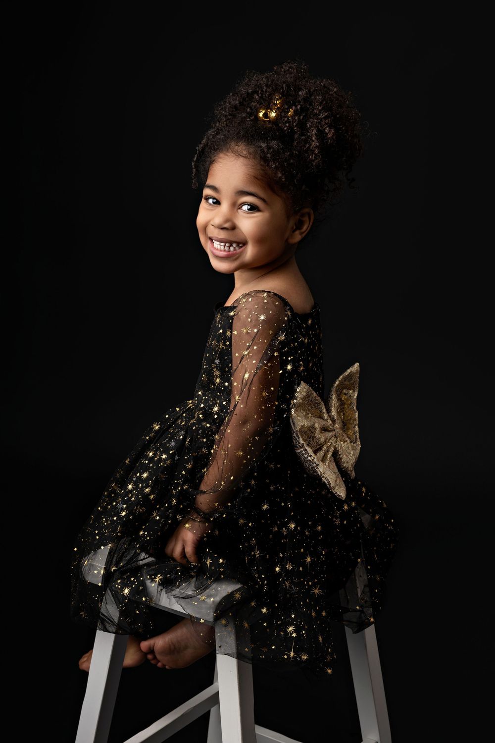 Young child in a sparkling black tulle dress with gold accents poses on a stool against dark background.