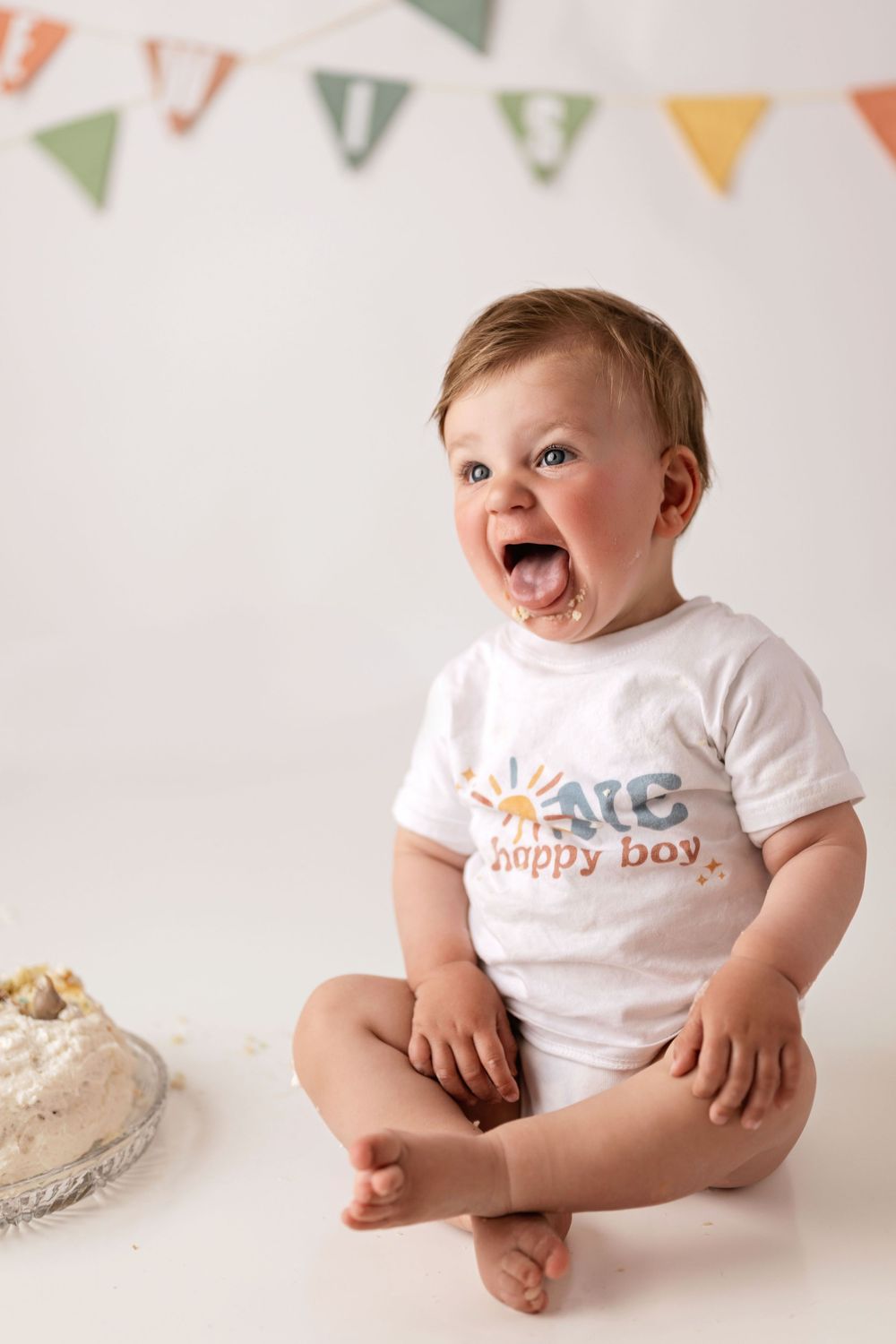 Baby wearing white 'happy boy' t-shirt sits on white backdrop with colorful triangle bunting decorations above.