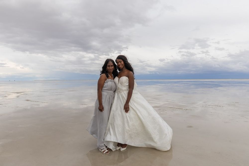 Two people in white dresses walk along a pristine beach shore under cloudy skies while holding hands.