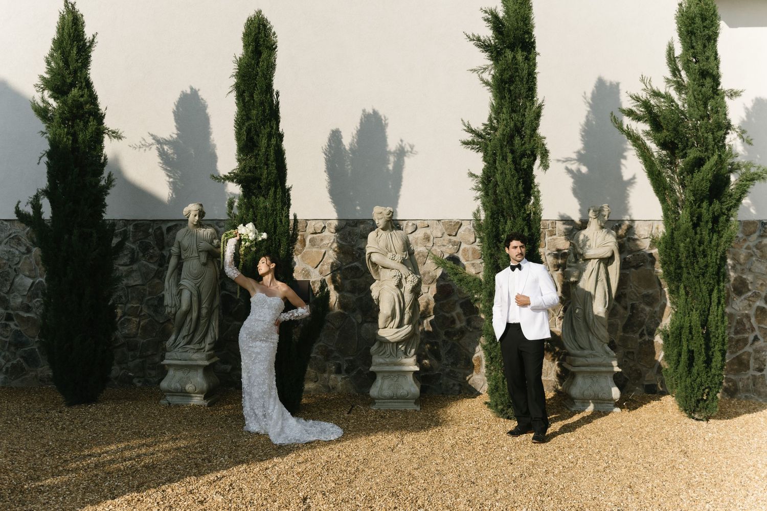 Elegant couple in formal attire posing among classical statues and cypress trees in a garden setting.