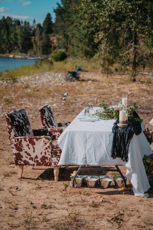 Rustic outdoor dining setup with white tablecloths laid across multiple tables on natural dirt terrain near forest scenery.