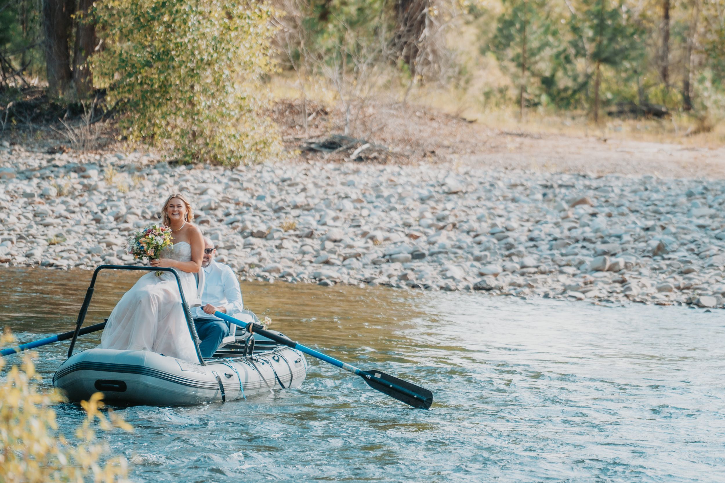 A person paddling a blue inflatable raft on a scenic river with rocky shoreline and trees in the background.