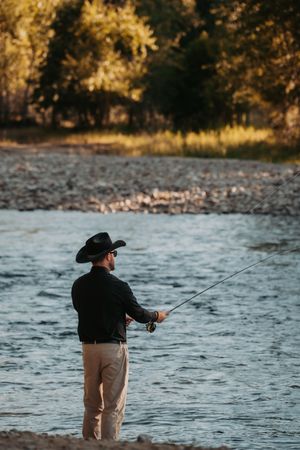 Person wearing a black outfit and cowboy hat fishing at the water's edge during golden hour.