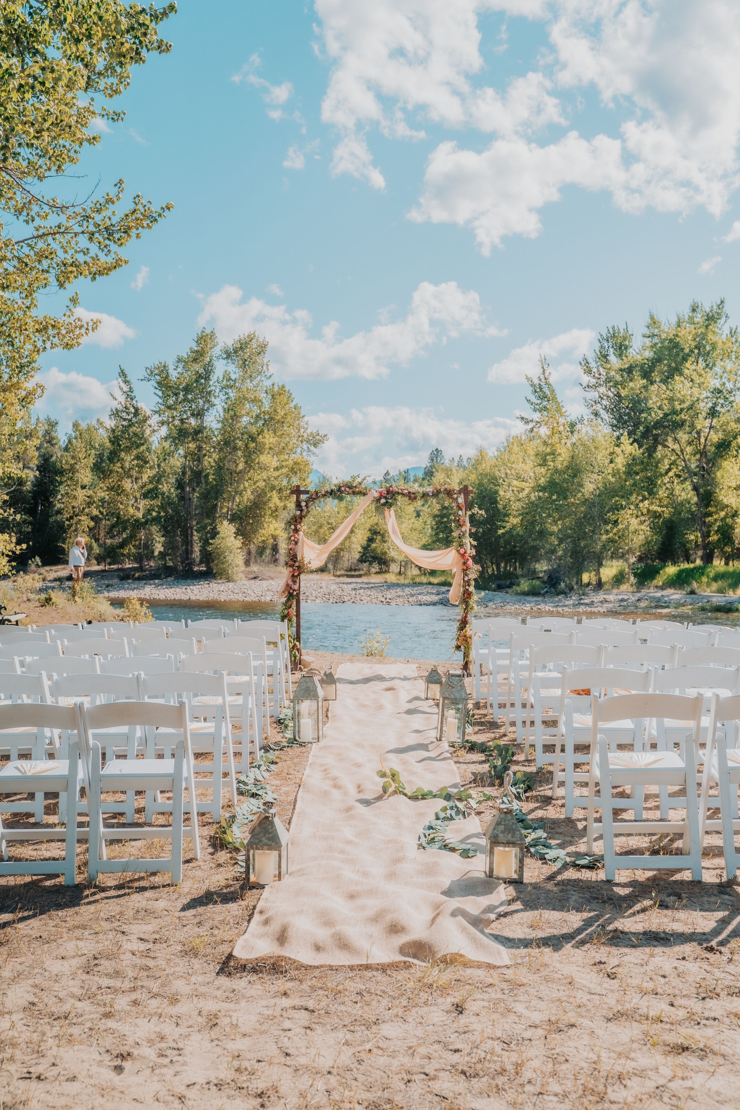 A rustic outdoor wedding ceremony setup with white chairs and floral arch by a river under blue skies and trees.