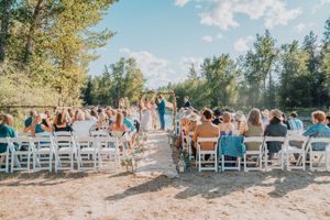 Wedding guests sit in white chairs on both sides of a sun-dappled outdoor aisle during a beautiful summer ceremony.