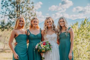 Bridal party in matching teal dresses stands together outdoors on sunny day with pine trees in background.