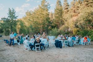 Wedding guests seated at tables with blue linens enjoy an outdoor reception surrounded by tall evergreen trees at sunset.